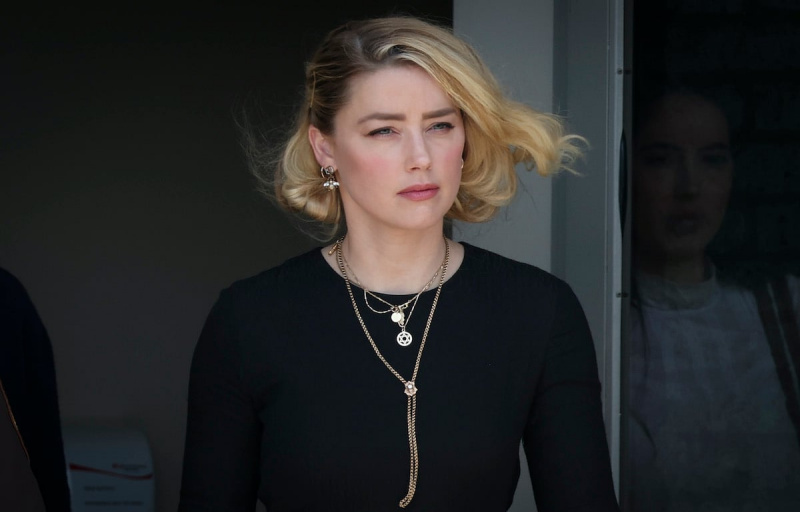  Amber Heard sale de un edificio y el viento le agita el cabello, vistiendo una camisa negra y múltiples collares.