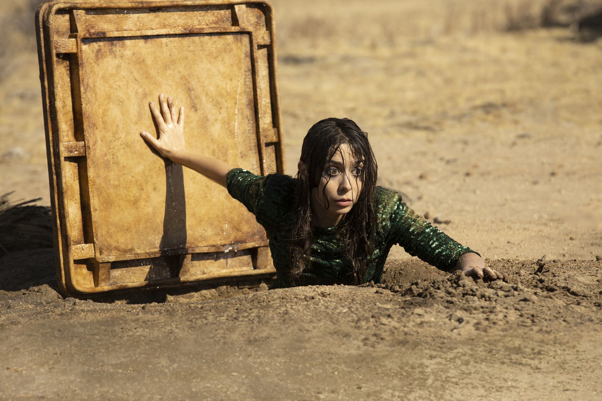 כריסטין מיליוטי בתפקיד לוז בתכנית לאהבה, מטפסת מתוך פתח המתכת אל נתיב מים תת קרקעי במדבר.