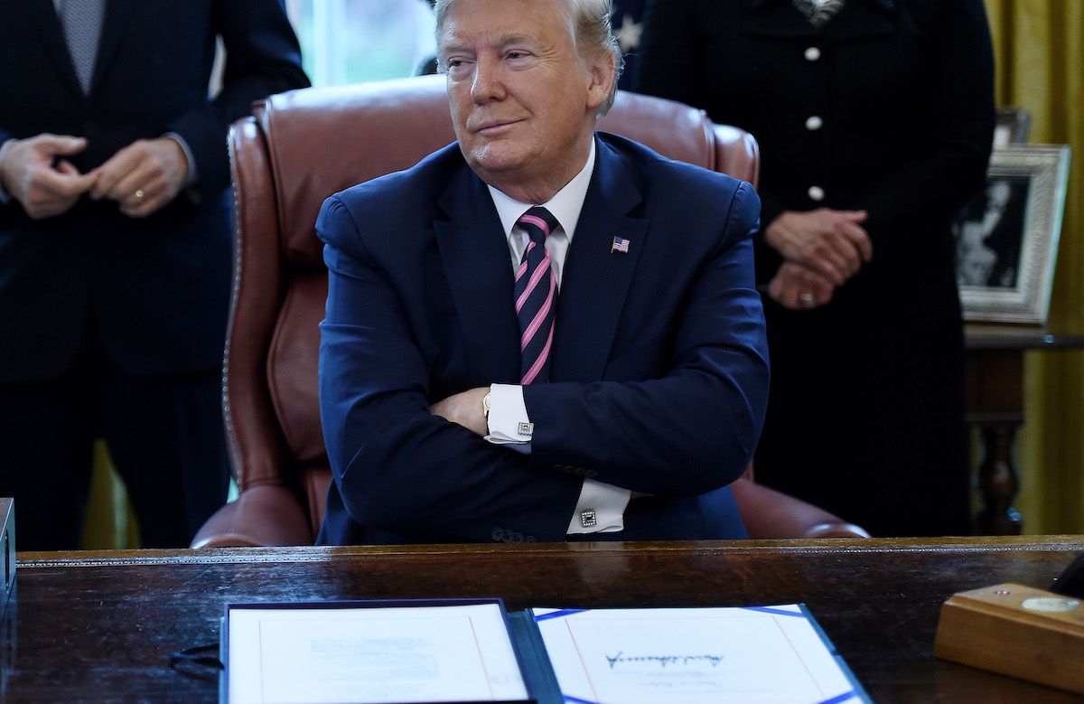 Donald Trump kollarını kavuşturuyor ve Oval Ofis masasının arkasında otururken kendini beğenmiş görünüyor.