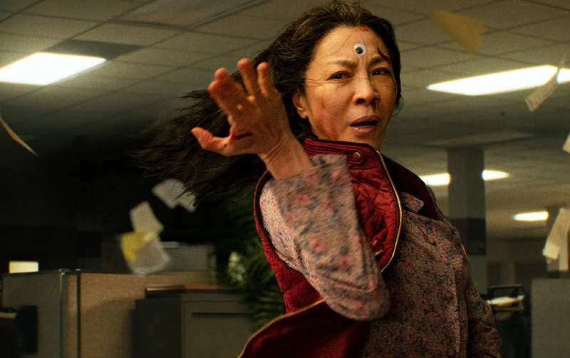   Evelyn (Michelle Yeoh) adopta una postura de lucha en una escena de'Everything Everywhere All at Once.'  She's standing in an office filled with cubicles and one of those googly eyes is attached to the center of her forehead. 