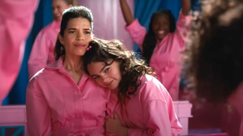   America Ferrera als Gloria und Ariana Greenblatt als Sasha in'Barbie.' Both are brown Latinas. Gloria is a woman with long, straightened dark brown hair styled half up-half down. Sasha's hair is also long and dark brown, but much fuller with waves, and held half-up half-down with a hair tie that's pink little balls. Both are wearing long-sleeved, pink jumpsuits as Sasha leans her head on Gloria's shoulder.