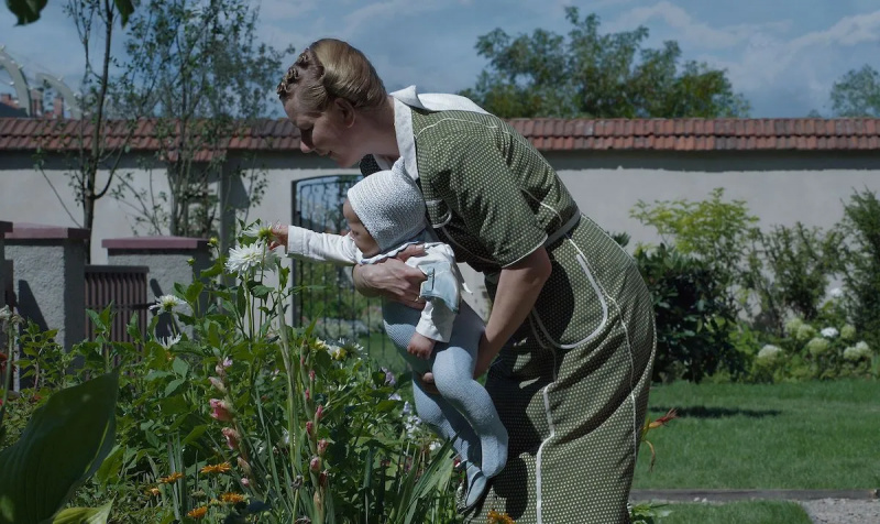   Hedviga sa skláňa nad kvetinovým záhonom a drží svoje dieťa. V rohu je viditeľná stena hraničiaca s Osvienčimom.