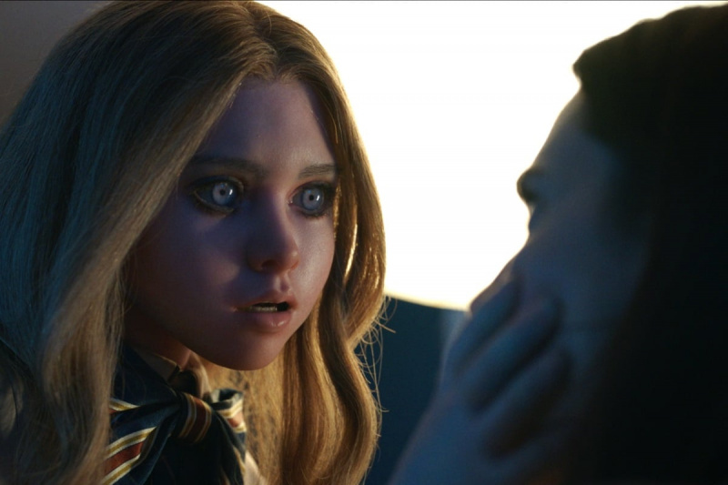   Slika lutke M3GAN (izgovarja se Megan) v istoimenskem filmu. Je lutka v naravni velikosti, ki je videti kot rdečelasko dekle s svetlimi očmi. Ona's leaning in menacingly while holding Allison Williams' face. 
