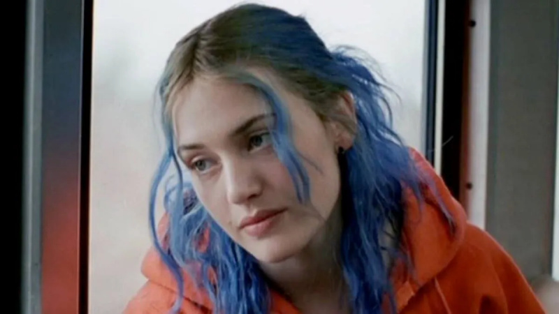   Slika Kate Winslet kot Clementine v'Eternal Sunshine of the Spotless Mind.' She is a white woman with dyed blue hair with light brown roots showing worn half up and half down. She's wearing a bright orange hoodie. 
