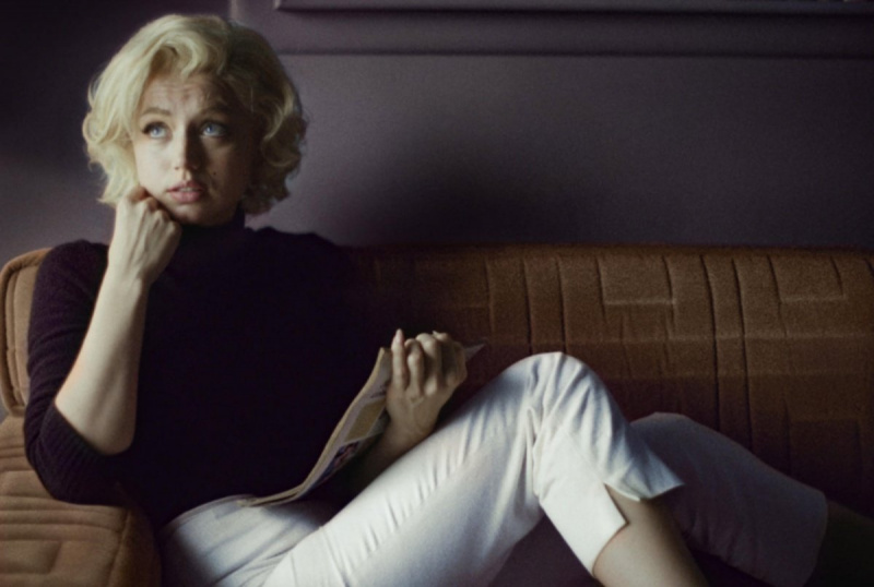   Ana de Armas, como Marilyn Monroe, está sentada em um sofá com um jornal na mão.