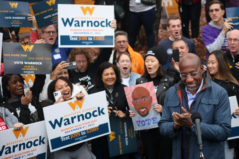  Raphael Warnock uśmiecha się i klaszcze, stojąc przed grupą kibiców trzymających transparenty kampanii.