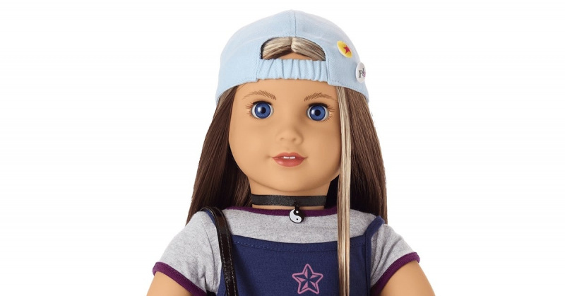  Un primer plano de una muñeca American Girl con una gorra de béisbol al revés, cabello castaño con una raya rubia en la cara y un collar yinyang.