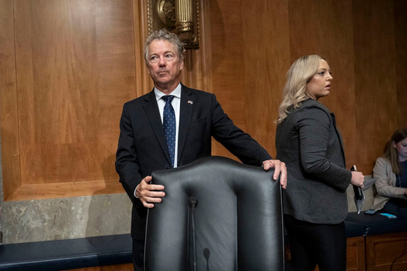   Rand Paul bir sandalyenin arkasında duruyor, gergin bir şekilde başka tarafa bakıyor.