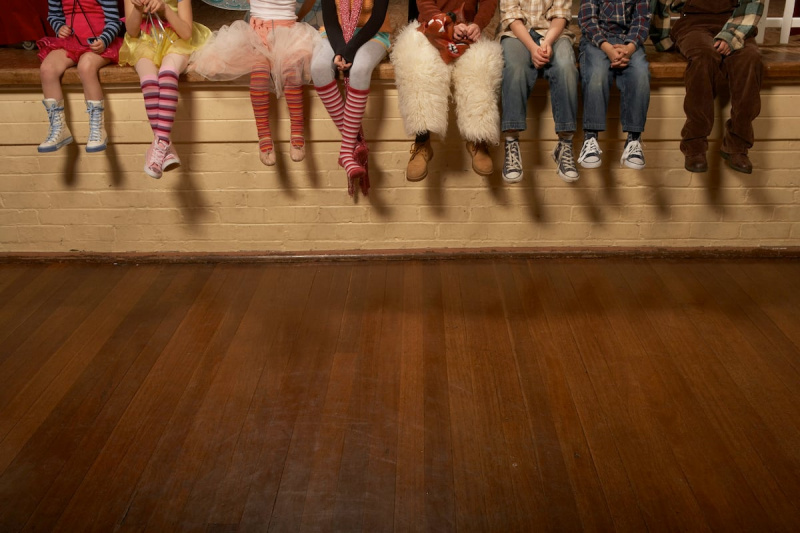   Vrsta mladih igralcev sedi na robu odra v različnih kostumih, gledano od pasu navzdol.