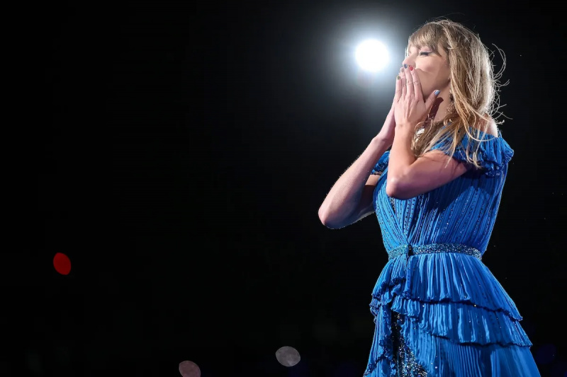  Taylor Swift v modrých šatech na jevišti vystupuje, drží si ruce u úst, aby políbila publikum.