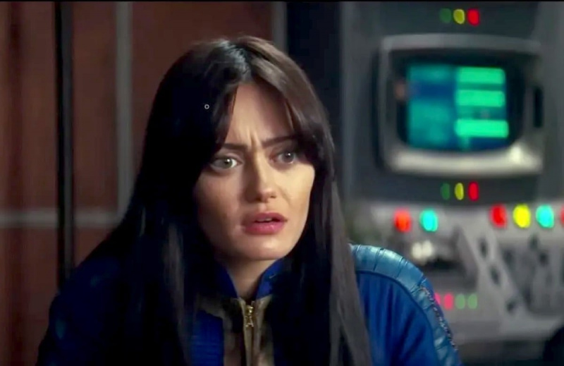   Ella Purnell como Lucy en una escena de Amazon's 'Fallout' series. She is a young white woman with long dark hair and long bangs parted down the middle. She's wearing a blue 'Fallout' vault suit and sitting in a room with computer consoles behind her. She has a confused expression on her face.