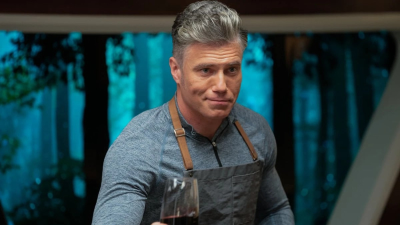   El Capitán Christopher Pike interpretado por Anson Mount en una escena de'Strange New Worlds.' He is a white man with mostly silver salt and pepper hair, wearing a blue, long-sleeved cotton zip-up shirt and an apron. He's holding out a glass of red wine with a smirk on his face.
