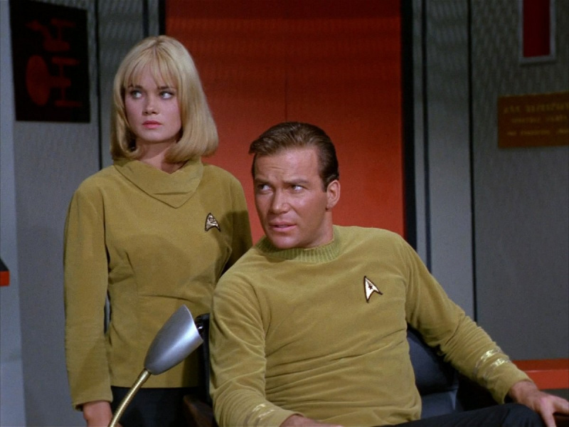  Yeoman Smith (Andrea Dromm) y el Capitán Kirk (William Shatner) en una escena de'Star Trek.' Smith is a blonde woman with a neck-length bob with bangs. She's wearing a gold Starfleet turtleneck and standing behind Kirk, a white man with brown hair wearing a gold Starfleet shirt and sitting in his captain's chair. They're both looking off camera.
