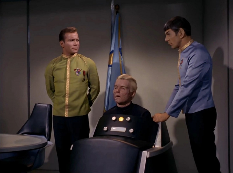   Kirk (William Shatner), Pike (Sean Kenney) y Spock (Leonard Nimoy) en una escena de'Star Trek.' Kirk is a white man with brown hair standing at ease while wearing a gold Starfleet dress uniform jacket and black pants. Pike is in a brainwave-operated wheelchair that he's encased in up to his chest. He's a white man with white hair and a burned face. Standing with his hand on the chair, Spock is a white Vulcan with pointy ears and short, dark hair wearing a blue Starfleet dress uniform jacket and black pants. 