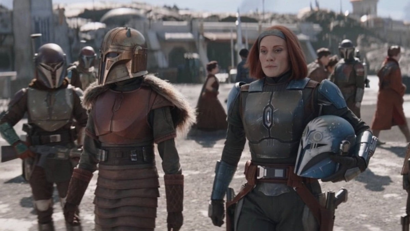   Emily Swallow como The Armorer y Katee Sackhoff como Bo-Katan en'The Mandalorian' on Disney+. They are walking through a busy area. The Armorer is in full Mandalorian armor, while Bo-Katan is in her armor, but has her helmet off and in one arm. Her face, grey headband, and chin-length red hair are visible. 