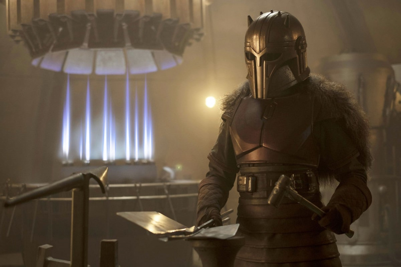   Emily Swallow como The Armorer en una escena de'The Mandalorian' on Disney+. She is in her full Mandalorian armor, and is holding a hammer, sheet metal, and tongs as she stands in the Forge. 