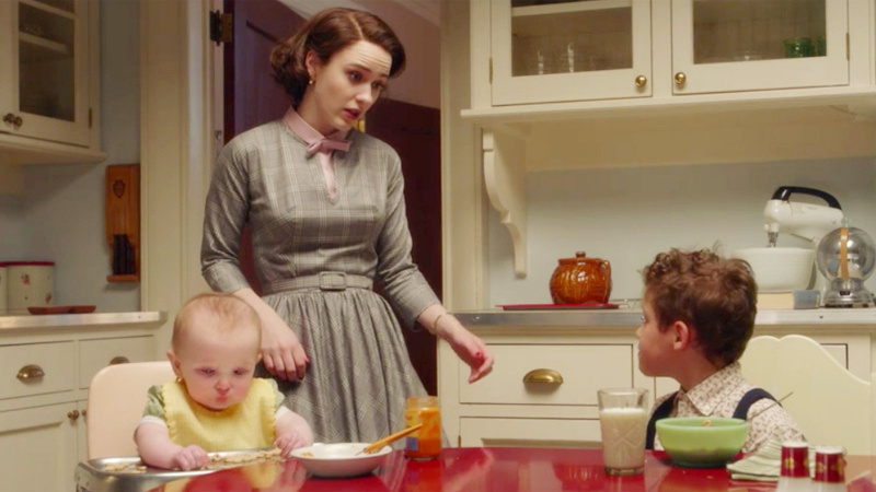   Rachel Brosnahan als Miriam „Midge“ Maisel mit Baby Esther und ihrem älteren Sohn Ethan in einer Amazon-Szene's 'The Marvelous Mrs. Maisel.' The kids are seated at the kitchen table as Midge stands talking to Ethan. She is a white woman with bobbed reddish-brownish hair and wearing a grey, belted, 1950's style dress with a pink collar and bow at her neck. 