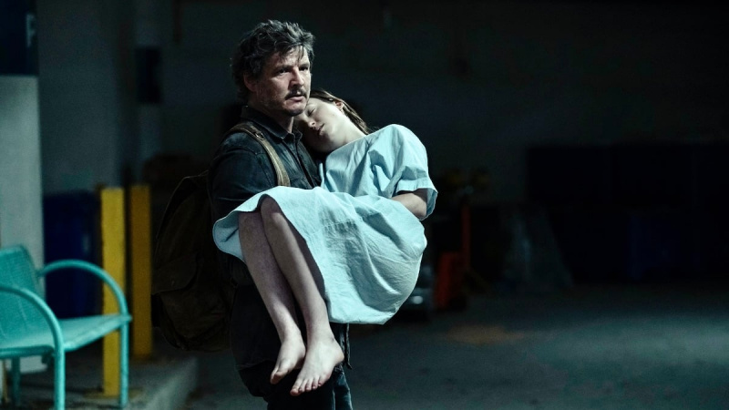   Obrázok Pedra Pascala (Joel) a Belly Ramsey (Ellie) v scéne z'The last of us' on HBO. Joel has dark, scruffy hair and a mustache and wears a dark jacket and a brown backpack. He's carrying Ellie, who is unconscious and in a hospital gown. He's walking through a parking garage.
