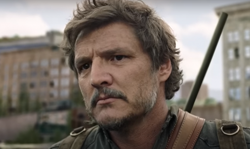   Obrázok Pedra Pascala ako Joela na'The last of Us' on HBO. Close-up image of Joel, who is outdoors and looking at something with a serious expression. His brown hair is shaggy, and he has a salt-and-pepper mustache and a light beard. He's wearing a rifle on his back and a green backpack and a greenish jacket. 