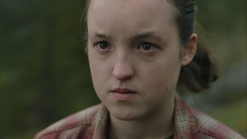  Obrázok Bella Ramsey ako Ellie na'The last of Us' on HBO. Close-up image of Ellie, who is outdoors and looking at something with a serious expression. Her dark hair is pulled back into a pony tail, and she's wearing a red plaid collared shirt. 