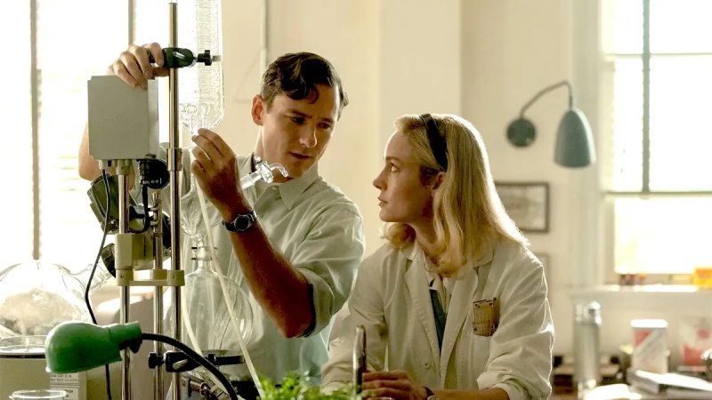   Bild von Lewis Pullman als Calvin und Brie Larson als Elizabeth in einer Szene aus'Lessons in Chemistry' on Apple TV+. Calvin is a white man with short brown hair wearing a white lab coat. Elizabeth is a white woman with shoulder-length blonde hair wearing a white lab coat. They are both standing at a counter in a chemistry lab looking at each other as Calvin adjusts a fluid bag on a stand.